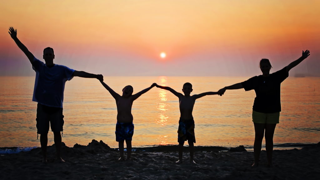 Família com pai, duas crianças de mesma idade e mãe, estão em pé com as mãos dadas e os braços abertos e o pôr do sol ao fundo